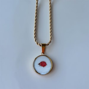 May include: Gold-toned necklace with a round pendant featuring a red ladybug design on a white background. The chain has a twisted rope design. The pendant is encased in a gold-colored frame.