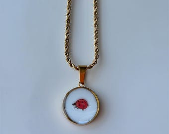 May include: Gold-toned necklace with a round pendant featuring a red ladybug design on a white background. The chain has a twisted rope design. The pendant is encased in a gold-colored frame.