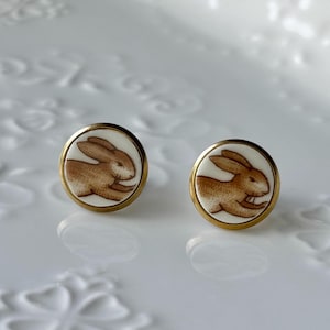May include: A pair of circular stud earrings featuring a white background with a brown rabbit illustration. Each earring is encased in a thin gold-toned metal frame, showcasing a detailed profile of a leaping rabbit in a consistent brown hue.