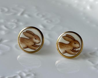 May include: A pair of circular stud earrings featuring a white background with a brown rabbit illustration. Each earring is encased in a thin gold-toned metal frame, showcasing a detailed profile of a leaping rabbit in a consistent brown hue.