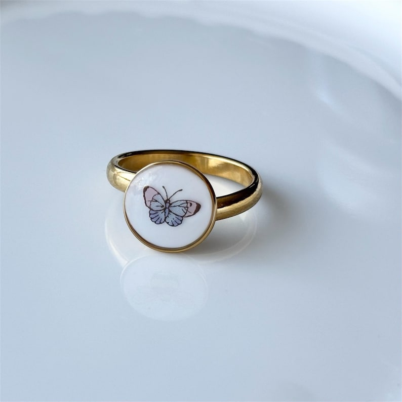 May include: A gold-colored ring with a round, white face featuring a delicate butterfly design in shades of pink, blue, and gray. The ring's band is smooth and polished, creating a contrast with the detailed butterfly image.