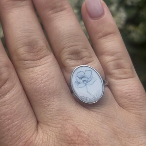 May include: An oval silver ring featuring a white inlay with a delicate, dark-lined botanical illustration of a single flower with petals and leaves centered on the face of the ring, set within a polished metallic band that encircles the oval design.