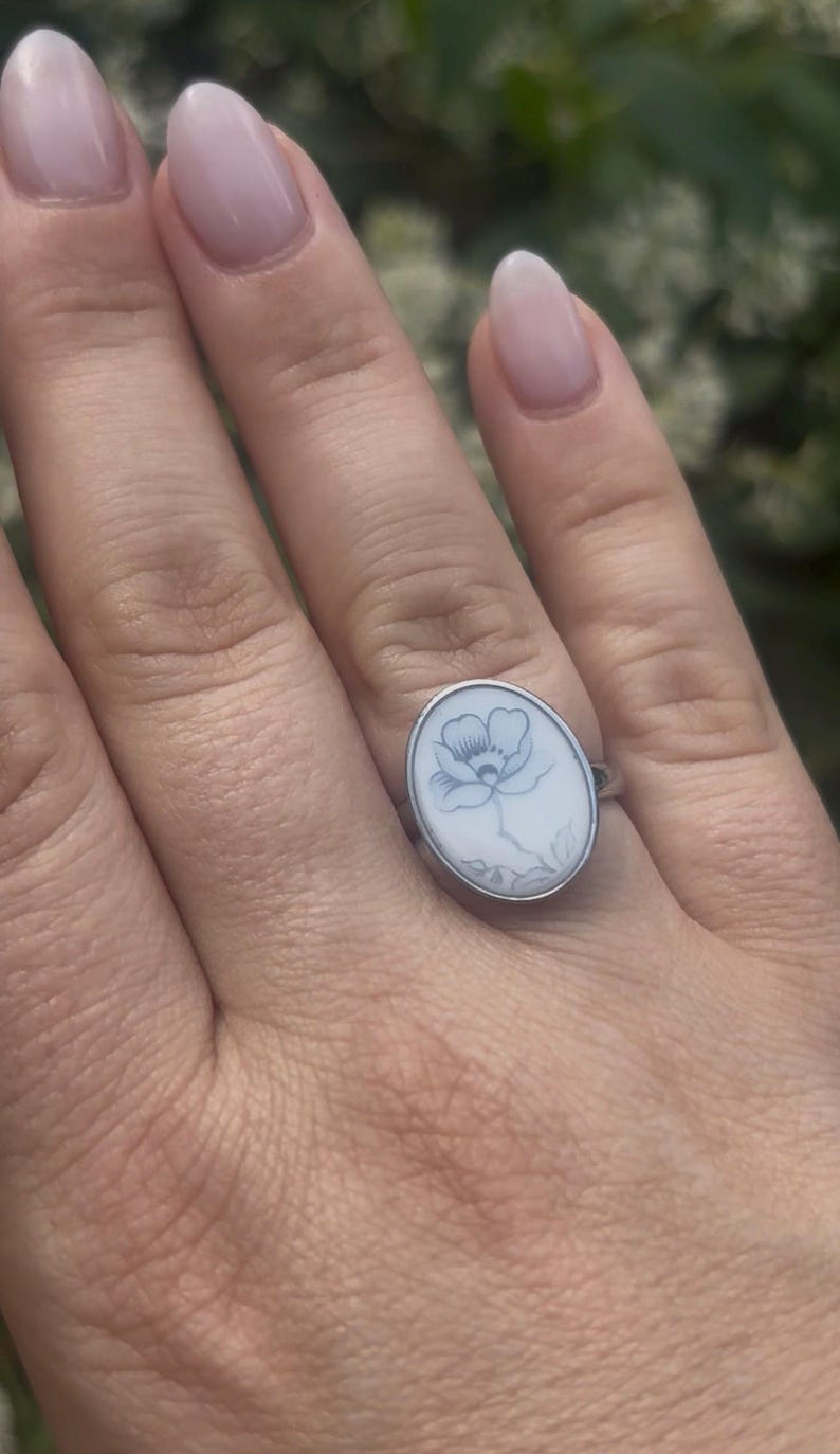 May include: An oval silver ring featuring a white inlay with a delicate, dark-lined botanical illustration of a single flower with petals and leaves centered on the face of the ring, set within a polished metallic band that encircles the oval design.