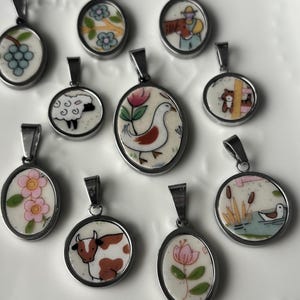 May include: A collection of oval and round pendants with silver-tone metal frames. Each pendant features a hand-painted design, including a sheep, cow, flowers, and a duck in a pond. The pendants are displayed on a white, textured surface.