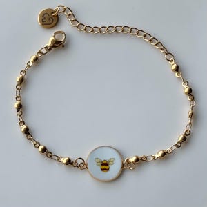 May include: Gold-toned bracelet featuring a round charm with a yellow and black bee design on a white background. The bracelet has a delicate chain and small gold beads. A small gold charm with a heart and the letter "B" is attached.