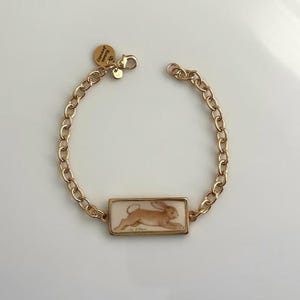 May include: A gold-toned chain bracelet featuring a rectangular pendant with a brown rabbit illustration. The bracelet includes a lobster claw clasp and a small round charm engraved with the text Arista Jewelry, creating a cohesive design.