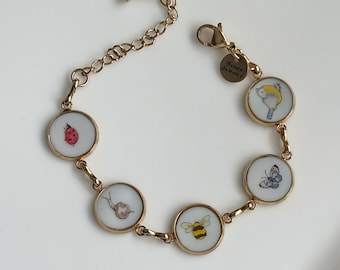 May include: A gold-toned chain bracelet featuring five circular charms with white backgrounds, each displaying a small illustration of a ladybug, snail, bee, butterfly, or bird. A small round tag attached near the lobster clasp reads Arista Jewelry.