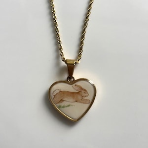 May include: A gold-toned rope chain necklace featuring a heart-shaped pendant. The pendant has a cream-coloured centre inlay displaying a brown rabbit illustration in a running pose over a small patch of green grass, framed by a polished gold border.