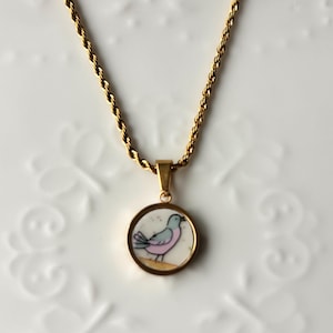 May include: A gold-toned rope chain necklace featuring a circular pendant. The pendant has a gold-toned metal rim and a central inlay depicting a small bird with a light blue head, a pink body, and dark outlines, standing on a tan line.
