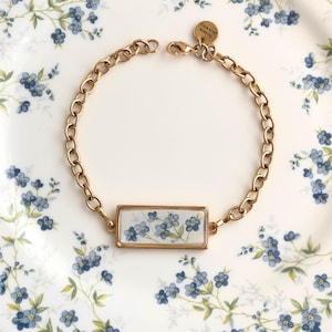 May include: A gold-toned chain bracelet featuring a rectangular pendant with a white background and a blue floral pattern. The bracelet includes a small circular charm engraved with the text Arista Jewelry and a lobster claw clasp closure.