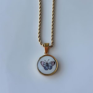 May include: Gold-toned necklace with a round pendant featuring a butterfly design. The butterfly has blue and pink details, set against a white background. The chain has a rope-like texture.
