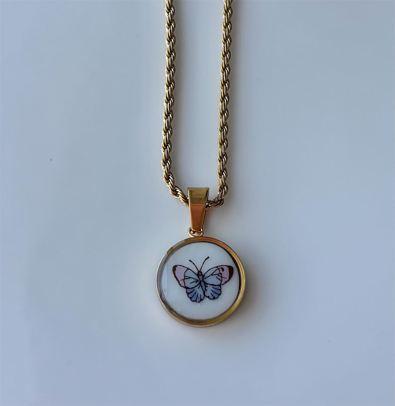 May include: Gold-toned necklace with a round pendant featuring a butterfly design. The butterfly has blue and pink details, set against a white background. The chain has a rope-like texture.