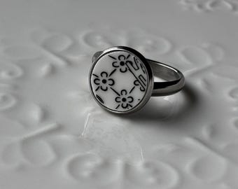 May include: A silver ring with a round, white face featuring a black floral pattern. The ring's band is a simple, polished silver. The floral design is composed of small, stylized flowers connected by lines.