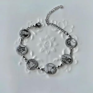 Könnte beinhalten: Silberfarbenes Armband mit runden Anhängern, die schwarz-weiße Illustrationen von Zeichentrickfiguren zeigen. Das Armband hat ein Kettendesign und einen Verschluss. Die Anhänger sind durch kleine silberne Glieder verbunden.