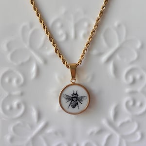 Puede incluir: Collar dorado con un colgante redondo con una ilustración detallada de una abeja en blanco y negro. El colgante tiene un fondo blanco y un borde dorado. La cadena es de diseño de cuerda trenzada.