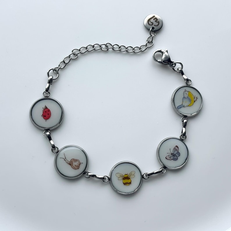 May include: Silver-toned bracelet featuring round charms with colorful illustrations of a ladybug, snail, bee, butterfly, and bird. The bracelet has a chain extension and a lobster clasp for secure wear.