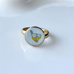 May include: A gold-colored ring with a circular, white face featuring a watercolor-style illustration of a small bird. The bird has blue, yellow, and gray coloring. The ring is set against a white background.