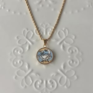 Peut inclure: Collier doré avec un pendentif rond représentant deux oies. Le pendentif a un fond bleu clair avec des accents jaunes et blancs. La chaîne a un design torsadé.