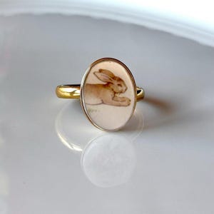Op de afbeelding: Een goudkleurige ring met een ovale zetting en een crèmekleurige inleg. De inleg toont een bruine illustratie van een rustend konijn. De ringband is glad en metaalachtig en reflecteert het licht op het gepolijste oppervlak.