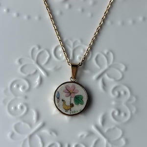 Peut inclure: Un collier à chaîne dorée avec un pendentif circulaire sur fond blanc. Le pendentif présente une illustration peinte à la main d'un petit oiseau jaune regardant une fleur rose avec des feuilles vertes et un petit accent bleu.