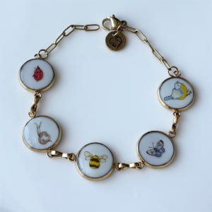 May include: Gold-toned bracelet featuring six circular charms with nature-themed illustrations. The charms depict a ladybug, snail, bee, butterfly, and a bird. The bracelet has a lobster clasp closure.