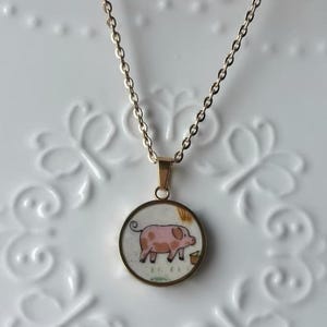 May include: Gold-toned necklace with a round pendant featuring a pink pig illustration. The pig is standing near a small brown bucket. The pendant has a gold-colored border and hangs from a delicate chain.