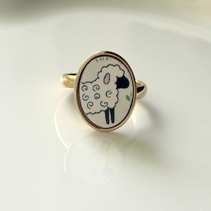 May include: A gold-toned ring featuring an oval-shaped setting with a white background. The center displays a graphic illustration of a sheep with curly white wool, a black face, and black legs. The sheep has a small pink ear and a green leaf.