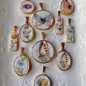 May include: A collection of gold-toned pendants with various designs, including butterflies, flowers, and a ladybug. The pendants are set on white backgrounds with gold rims. The shapes vary from round to oval and rectangular.