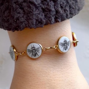 May include: A gold-toned chain bracelet featuring multiple circular charms. Each charm contains a white inlay with a detailed black line drawing of a bee. The circular charms are connected by small gold-toned metal links in a repeating pattern.