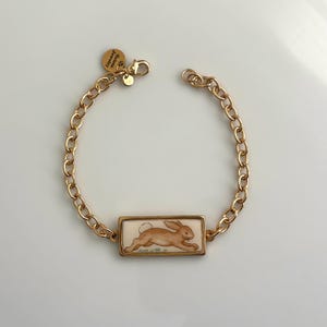 Puede incluir: Una pulsera de cadena en tono dorado con un colgante rectangular que presenta una ilustración pintada de un conejo marrón. La pulsera incluye un pequeño dije circular grabado con el texto Arista Jewelry, unido cerca del cierre de mosquetón.