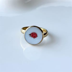 May include: A gold-colored ring with a circular face featuring a red and black ladybug design on a white background. The ring has a simple, elegant band.