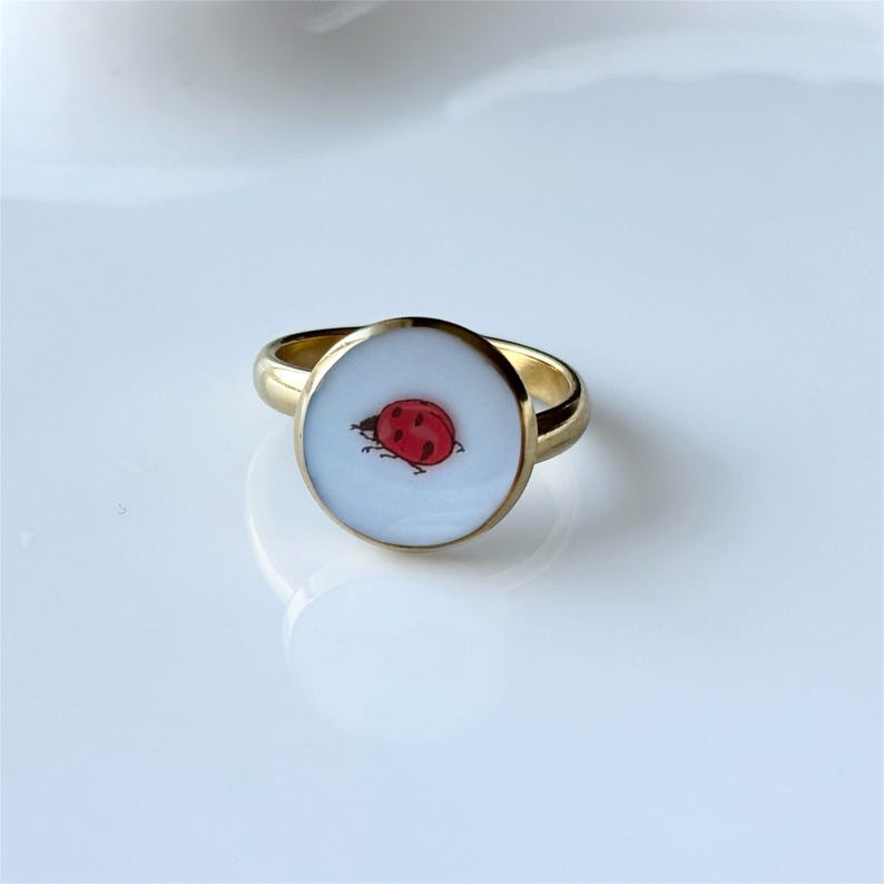 May include: A gold-colored ring with a circular face featuring a red and black ladybug design on a white background. The ring has a simple, elegant band.
