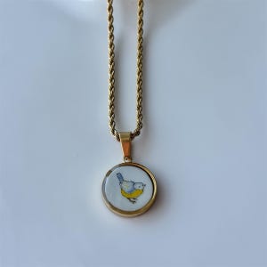 May include: Gold-toned necklace with a round pendant featuring a blue and yellow bird illustration on a white background. The pendant is framed in gold and hangs from a twisted rope chain.