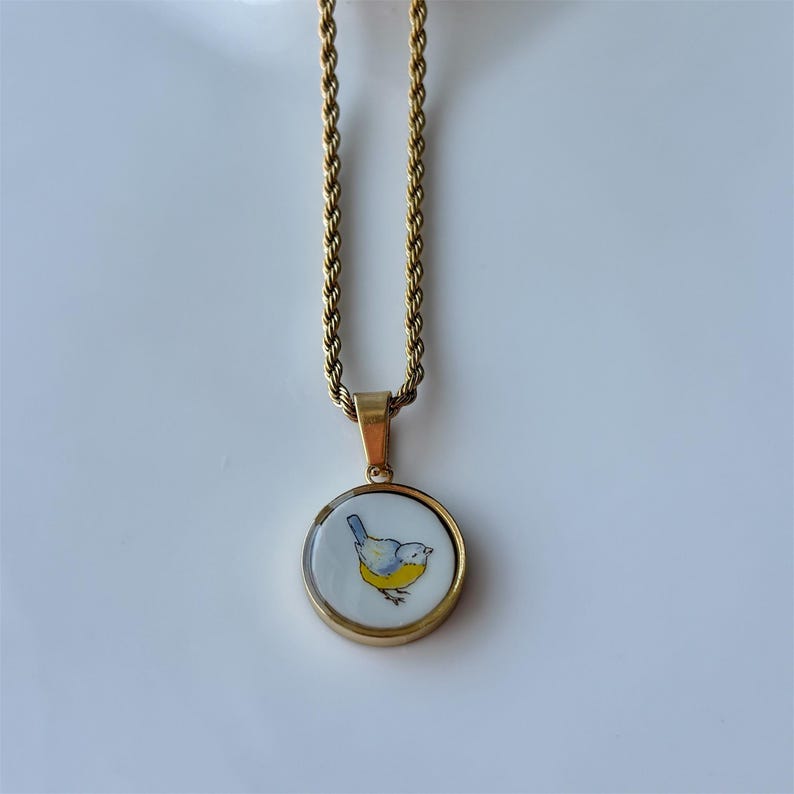 May include: Gold-toned necklace with a round pendant featuring a blue and yellow bird illustration on a white background. The pendant is framed in gold and hangs from a twisted rope chain.
