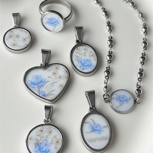 May include: A collection of jewelry pieces featuring a floral design. The set includes pendants in round, oval, and heart shapes, a ring, and a necklace. Each piece has a silver-tone metal frame and a white background with blue and yellow floral patterns.