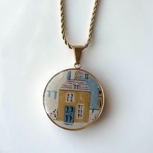 Könnte beinhalten: Goldfarbene Halskette mit rundem Anhänger, der eine Miniaturhaus-Illustration zeigt. Das Haus ist gelb mit blauer Tür und Fenstern, vor hellblauem und weißem Hintergrund. Die Kette hat ein gedrehtes Seildesign.