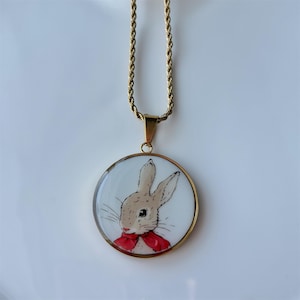 May include: Gold-tone necklace featuring a round pendant with a bunny illustration. The bunny has tan fur and a red bow tie. The pendant is set against a white background.