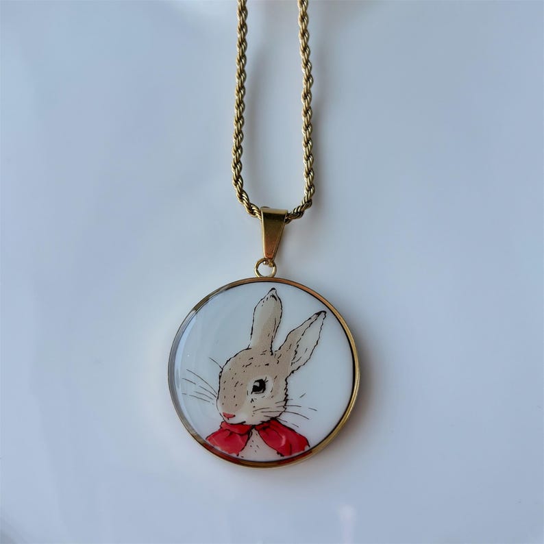 May include: Gold-tone necklace featuring a round pendant with a bunny illustration. The bunny has tan fur and a red bow tie. The pendant is set against a white background.
