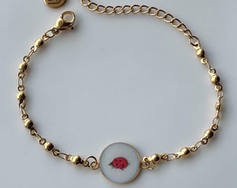 May include: Gold-toned bracelet featuring a central circular charm with a red ladybug design. The bracelet has a delicate chain with small, round beads and a lobster clasp closure. A small, round charm with the initials "G3" is also present.