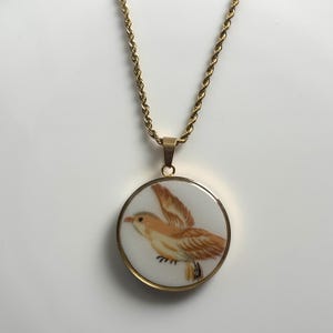 May include: A gold-toned rope chain necklace featuring a circular pendant. The pendant has a white background with a painted illustration of a light brown bird in flight with its wings spread. The metal frame of the pendant is gold-toned.