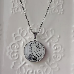 Peut inclure: Un collier argenté avec un pendentif rond représentant Winnie l'Ourson et Porcinet en noir et blanc. Le pendentif a un fond quadrillé et un cadre argenté. La chaîne est torsadée.