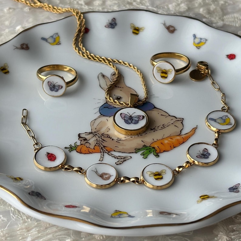 May include: A gold-toned jewelry set featuring a necklace, two rings, and a bracelet. Each piece has a white circular pendant with a nature-themed design, including butterflies, bees, and a ladybug. The jewelry is displayed on a decorative plate with a bunny illustration.