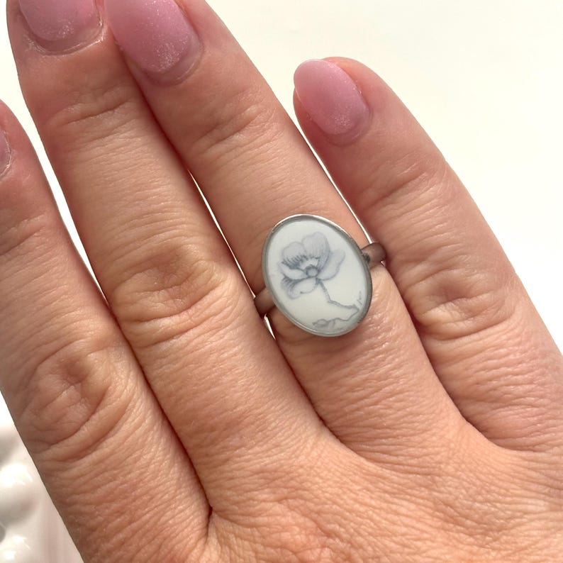 May include: A silver-toned ring featuring an oval-shaped setting with a white background. The center of the oval displays a delicate, hand-drawn illustration of a single flower in shades of grey, showcasing detailed petals and a stem.