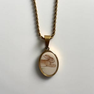 Peut inclure: Un collier à chaîne torsadée dorée avec un pendentif ovale. Le pendentif possède un cadre en métal doré poli entourant une incrustation de couleur crème qui présente une illustration détaillée d'un lapin brun couché sur le sol.
