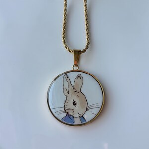 May include: A gold-toned necklace featuring a round pendant with a cartoon rabbit illustration. The rabbit is depicted in a blue jacket against a white background. The chain has a twisted rope design.