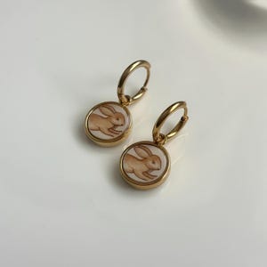 May include: A pair of gold-toned hoop earrings, each featuring a circular pendant with a white background and a brown illustration of a rabbit in a leaping position. The metal hoops are polished and circular, connecting to the round charms.