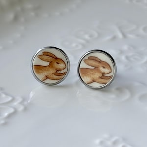 May include: A pair of round stud earrings featuring a cream-colored background with a brown illustration of a rabbit in profile. Each circular earring is encased in a thin silver-toned metal frame, showcasing a detailed bunny design.