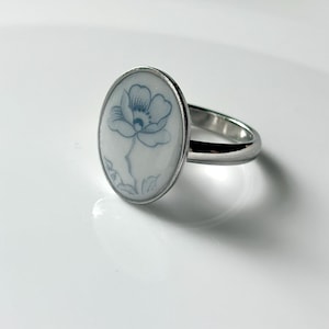 May include: A silver-toned ring featuring an oval-shaped white ceramic inlay. The inlay is decorated with a delicate, hand-painted blue floral illustration of a single flower with petals and a stem, set within a polished metal band.