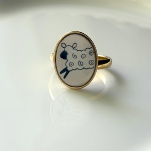 May include: A gold-toned ring featuring an oval-shaped face with a white background. The center displays a stylized illustration of a sheep with a black head, black legs, and a white, curly wool body outlined in black with small pink ears.
