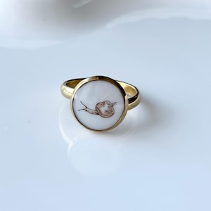 May include: A gold ring with a circular white face, showcasing a brown snail illustration. The ring has a polished finish and is set against a white backdrop. The snail is depicted in a simple, illustrative style.